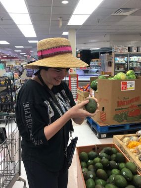 Shopping \ Angelman Syndrome News \ Columnist Mary Kay's daughter, Jessie, dressed in a beige and pink summer hat, smiles while inspecting an avocado at a supermarket.