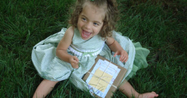 Angelman child's happy demeanor | Angelman Syndrome News | Juliana sits on the grass in a pretty green dress with a green bow on her head while flashing a smile. She is opening a gift.