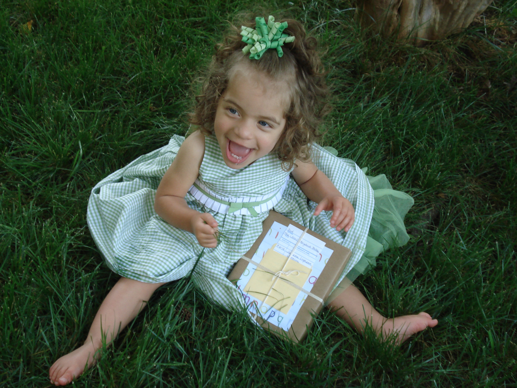 Angelman child's happy demeanor | Angelman Syndrome News | Juliana sits on the grass in a pretty green dress with a green bow on her head while flashing a smile. She is opening a gift.