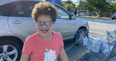 Outings with Angelman | Angelman Syndrome News | With a bright pink shirt and the sun behind her, Juliana smiles in a shopping center parking lot on an outing with her mom.