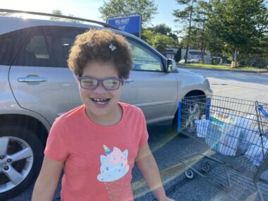Outings with Angelman | Angelman Syndrome News | With a bright pink shirt and the sun behind her, Juliana smiles in a shopping center parking lot on an outing with her mom.