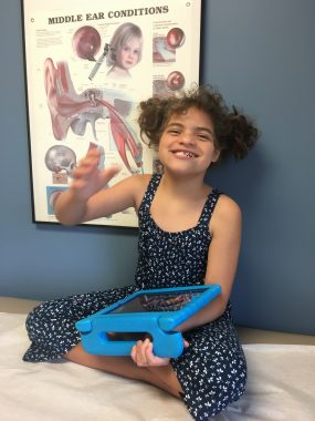 Angelman and reflux | Angelman Syndrome News | Juliana smiles and waves while sitting cross-legged in the pediatricians office
