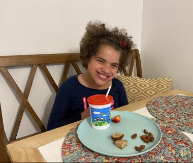 Angelman and reflux | Angelman Syndrome News | Juliana flashes a broad smile while sitting at the dinner table eating a healthy snack