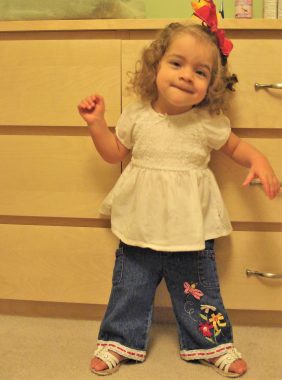 Learning to walk with Angelman | Angelman Syndrome News | A 2-year-old Juliana stands up with the support of a dresser. She wears a cute red bow in her curly hair.