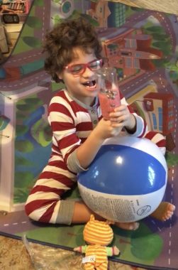 holiday gifts with Angelman | Angelman Syndrome News | Juliana wears red and white striped pajamas and sits on a carpet holding a beach ball and a homemade shaker on Christmas morning.