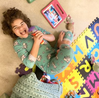 sick | Angelman Syndrome News | Juliana lies on her back on the floor in her room. She is wearing pajamas and laughing, and is surrounded by toys.