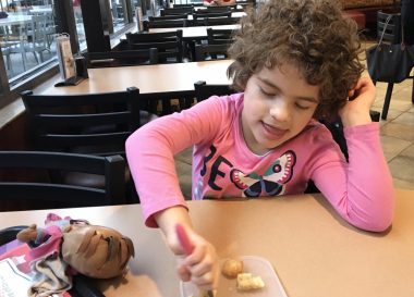 toy | Angelman Syndrome News | Juliana sits at a table in a restaurant and eats, with Ladybug perched on the table right next to her.