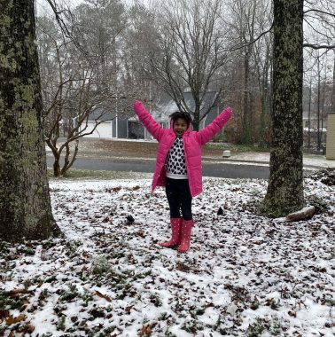 snow day | Angelman Syndrome News | Jessa stands outside in the snow wearing a pink jacket and boots. She's laughing and reaching her arms toward the sky.