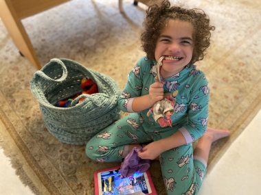 snow day | Angelman Syndrome News | Juliana smiles and sits on the floor in her pajamas with her doll, toys, and iPad