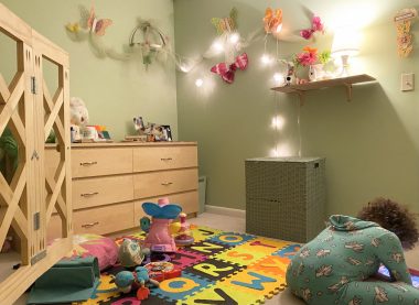 realistic self-care | Angelman Syndrome News | Sabrina snaps a photo of her daughter's bedroom while Juliana sits on the floor with her iPad. The light green room features twinkling lights, butterflies on the wall, a foam alphabet mat, and lots of toys.