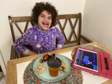 Angelman behaviors | Angelman Syndrome News | Juliana smiles as she sits in her purple pajamas at a table in front of her breakfast. Also on the table is her red iPad/talker.