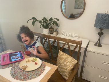 parent challenges of angelman syndrome | Angelman Syndrome News | Juliana smiles while sitting at the breakfast table, with a plate of food and her pink tablet.