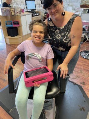 getting a haircut | Angelman Syndrome News | photo of Juliana in the stylist's chair looking at the camera, with the stylist behind her