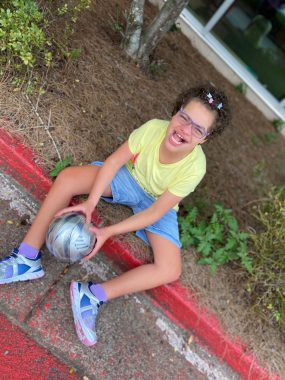 making exercise a priority | Angelman Syndrome News | a photo of Juliana, in yellow shirt and blue shorts, sitting by a plant bed, with a ball in her hands
