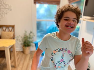 benefits of music therapy | Angelman Syndrome News | Juliana is dancing and smiling. She's wear a light blue T-shirt with a floral peace sign