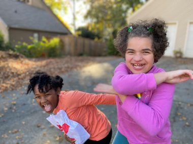 Two girls laugh and celebrate in the driveway of a house, with leaves covering the ground indicating it is autumn. One girl holds a bag of Chick-fil-A food. One is wearing an orange shirt and one a pink shirt. The girl on the right has Angelman syndrome.