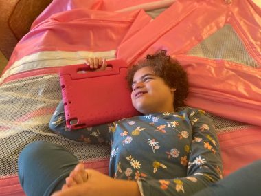 A girl lies on a red travel safety bed and holds an augmentative and alternative communication device. She looks to her right while holding her foot. 