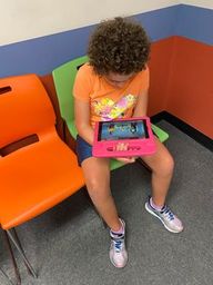 Juliana sits on a green chair at the doctor's office looking down at her iPad. Her orange T-shirt matches the otherwise orange furniture.
