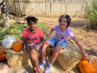 Two girls sit on bales of hay with pumpkins in an outdoor yard. They're both wearing shorts, and it's sunny out. The hay and pumpkins indicate it's fall.
