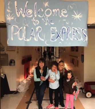 Two teachers stand with two young girls in a school hallway. Above them hangs a large, light blue banner that reads "Welcome to the Polar Express" in white and is surrounded by snowflakes. In the background are Christmas lights and other decorations.