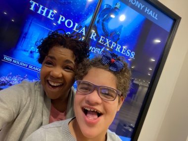 A woman and her daughter smile for a selfie in front of a poster for "The Polar Express" at the Atlanta Symphony Hall. The girl, who has Angelman syndrome, is wearing a gray sweater, glasses, and a blue and red bow in her hair. The woman is wearing a pink shirt and gray cardigan.