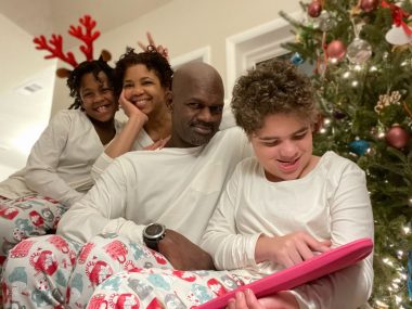 A family of four sits in front of their Christmas tree wearing matching pajamas, which include a white long-sleeved shirt and colorful patterned pants. Sabrina and her husband sit in the middle. Their younger daughter, Jessa, is on the left and wearing red reindeer antlers. Their older daughter, Juliana, who has Angelman syndrome, is on the right and smiling at a pink tablet she's holding.