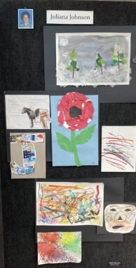 A black gallery wall features several pieces of art by a young girl, whose name, Juliana Johnson, and picture are posted at the top. The art includes some colorful scribbles, a paper mosaic of a red flower, a collage in the shape of a "J," and a painting of evergreen trees against a gray background.