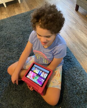 A 13-year-old girl with Angelman syndrom sits cross-legged on a blue rug on a wooden floor. The camera angle is looking down on her, showing the front of her and the top of her head. She's smiling, looking down, and holding a child's red iPad on her lap.