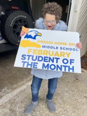 A girl stands in a driveway just outside of a garage holding a sign that reads "Proud home of middle school February student of the month" in blue and yellow letters. She has short brown curly hair and glasses and wears a light blue puffy jacket, jeans, and gray boots.