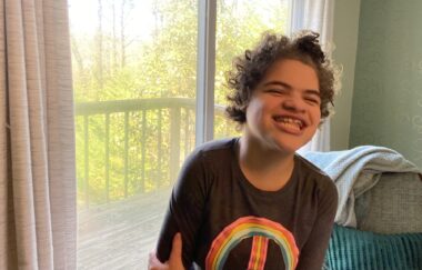A teenage girl with short, brown, curly hair smiles while dancing in her living room. She's wearing a brown T-shirt with what appears to be a rainbow peace sign, and behind her are floor-length windows looking out on a wooden deck.