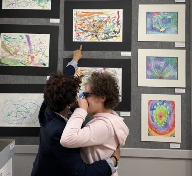 A woman, her back to the camera and wearing dark blue clothing, points to what appears to be crayon artwork just above her, part of a display of many such pieces of art. She is embracing a girl in a pink shirt with hoodie and a pair of glasses. They appear to be the same height.
