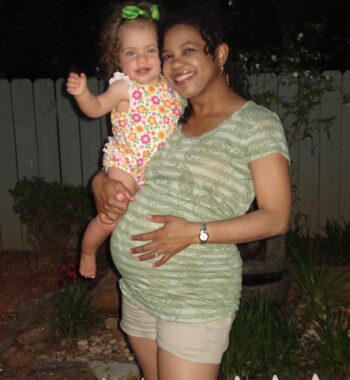 A visibly pregnant woman stands outside holding her young daughter with her right arm. Her left hand is resting on her belly. It's night, and they appear to be in their backyard. 
