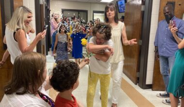 A school hallway shows a fifth-grade parade at the end of the year. A girl wearing yellow pants and a white shirt walks in the parade and is showered with confetti. Teachers stand to the side with smiles, taking photos on their cameras.
