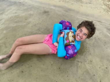 A girl reclines on solid beach sand, with her grinning face toward the camera. She wears a bright blue shirt and pink underpants, with what appear to be largely purple flotation devices on each arm.