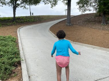 A girl wearing a bright blue shirt and pink underpants stands with her back to the camera on a concrete gray path, with gray raised borders on either side. The path has a slight incline for several yards, then curves slightly right and has a steeper incline. On either side of the path there is brown ground, with some low, green shrubbery and a couple of trees.