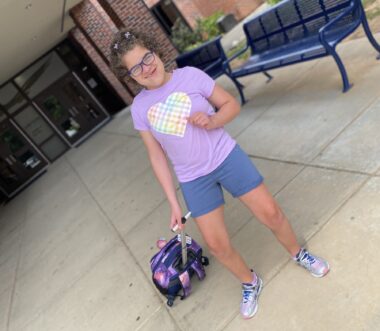 A girl strikes a pose in front of what appears to be a middle school. She has short, curly brown hair and glasses and is wearing blue shorts and a purple shirt with a rainbow heart on it. With her right hand, she's pulling a pink and purple roller backpack behind her. 
