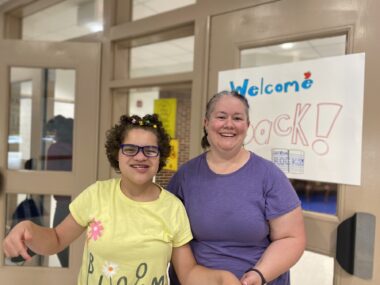 A 13-year-old girl smiles for a photo with her teacher. The girl is wearing a bright yellow T-shirt with pink and white flowers that reads "Bloom," and the teacher is wearing a purple shirt. They appear to be standing in a hallway at school in front of a handmade sign that reads "Welcome back!"