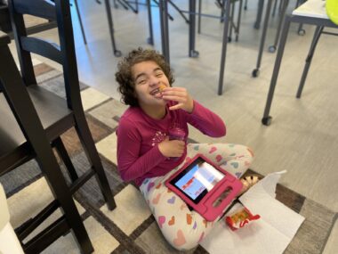 A 13-year-old girl sits cross-legged on a carpet in her mom's classroom. There's a tall chair to her right, and what appears to be several tables to her left. She's wearing a long-sleeve pink shirt and white leggings with pink and purple hearts. A pink iPad rests on her lap, and there's a small box of Chick-fil-A chicken nuggets on a paper towel on the floor in front of her. She's happily eating one as her mom snaps a photo.