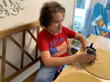 A 13-year-old girl with curly hair and a red T-shirt with a U.S. flag sits at a small breakfast table clutching a cup in both hands. She smiles and stares satisfactorily at the cup. 