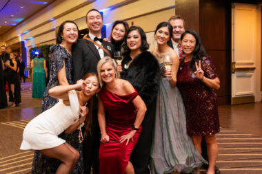 A group of nine adults pose for a photo at an Angelman syndrome gala. All are dressed in formal attire and smiling.