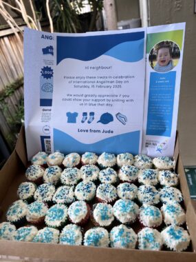 A large box containing about 40 mini cupcakes is propped open on what appears to be a table outside. The cupcakes all have white frosting and blue sprinkles. Behind them, several sheets of paper offer information about the author's son and Angelman syndrome. The paper in front has a message that reads, "Hi neighbour! Please enjoy these sweets in celebration of International Angelman Day on Saturday, 15 February, 2025. We would greatly appreciate if you could show your support by uniting with us in blue that day. Love from Jude."