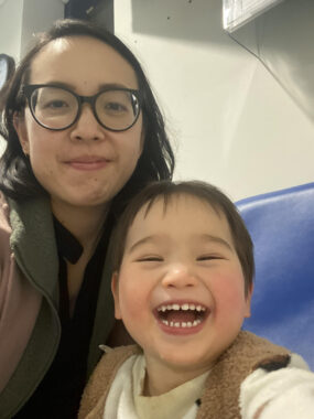 A woman takes a selfie with her 2-year-old son while sitting in the emergency room. She is giving a weary smile while the boy is laughing happily.