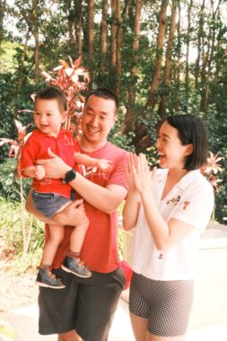 A man stands holding his 3-year-old son while his wife stands next to him and smiles at the boy. They are standing outside in some sort of lush environment.