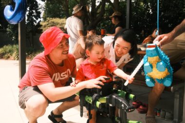 A 3-year-old boy holds out a small stick to hit a piñata. He's using some sort of mobility aid to help support him as he stands, and his parents are crouched beside him.