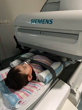 A young boy is strapped to a machine in the hospital. His body is inserted into the machine halfway, and he has earplugs and something in his mouth. 