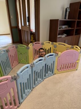 A young boy sits within a perimeter of blue, pink, and yellow plastic child barriers on the floor of a living room.