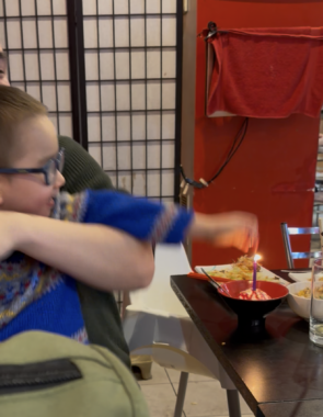 A blurry phone photo of a young child in glasses sitting on someone's lap and quickly reaching to his left to grab a burning birthday candle. 