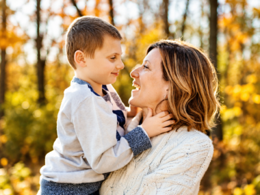 A woman holds a young boy with lots of autumn-leafed trees behind them.
