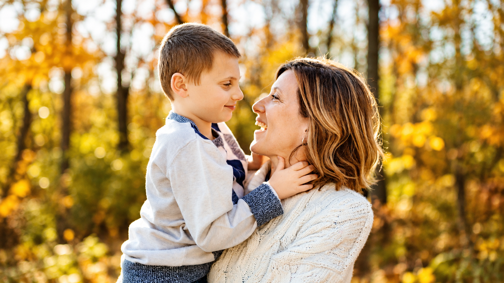A woman holds a young boy with lots of autumn-leafed trees behind them.